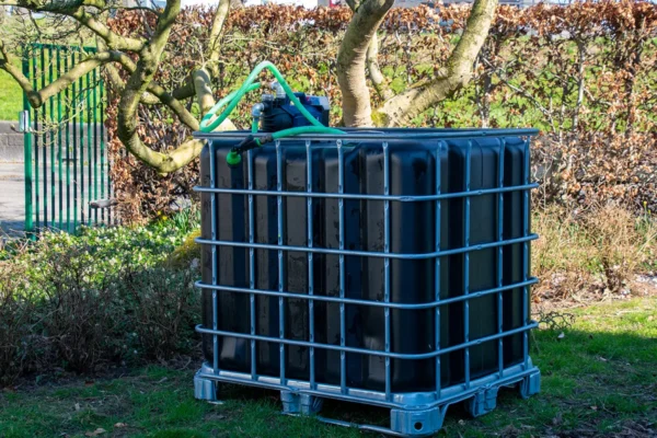 cuve-ibc-noir-avec-pompe-sur-batterie-1000L-renouvele-palette-acier-plastique-vue-de-cote-arrieere-avec-tuyau-sortant-dans-environnement-de-jardin.webp